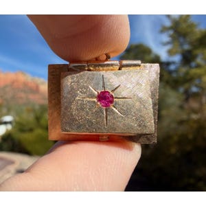 May include: A gold-toned, rectangular jewelry box with a textured surface. The box features a starburst design around a central, round, red gemstone. The box is held between two fingers, with a blurred outdoor background.