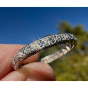 May include: A silver ring with a series of small, clear gemstones set around the band. The ring is held up against a bright blue sky and green foliage, highlighting the sparkle of the stones. The ring appears to be vintage or antique.