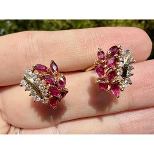May include: A pair of gold-toned earrings featuring a cluster of marquise-cut, deep red gemstones and small, clear gemstones. The earrings have a leaf-like design, with the red stones forming the leaves and the clear stones adding sparkle.