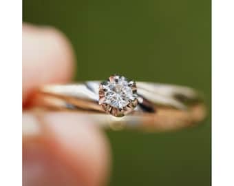 May include: A gold and silver ring with a clear diamond, shown in close-up. The ring is held against a blurred green backdrop, emphasising the diamond's brilliance and the ring's design. The band appears to be a mix of gold and silver.