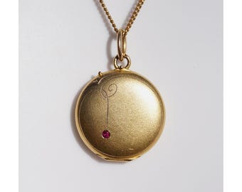 May include: A gold-colored locket pendant with a delicate chain. The round locket features a small red gemstone and an engraved design. The jewelry is displayed against a white background, highlighting the pendant's details.