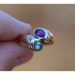 May include: A gold and silver ring with two oval gemstones, one purple and one blue. The ring is decorated with small, clear accent stones. The ring is held up by a hand.