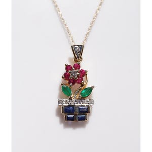 以下が含まれることがあります： 植木鉢に入った花の形をしたゴールドのペンダントネックレス。花には赤いルビーの花びらと中央に透明な石があしらわれています。茎からは2枚の緑色のエメラルドの葉が伸びています。鉢は青いサファイアのバゲットカットと透明な石で装飾されています。