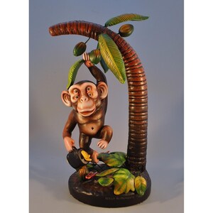May include: A brown monkey sculpture with four eyes hangs from a brown palm tree with green leaves and coconuts. The monkey holds a black object with a yellow flower. The base is dark brown with green leaves and the artist's signature.