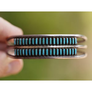 Puede incluir: Brazalete de plata con dos filas de piedras turquesas. Las piedras turquesas son rectangulares y están engastadas en una banda de plata. El brazalete se muestra sobre un fondo verde borroso.