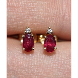 May include: A pair of stud earrings featuring oval-cut deep red rubies set in yellow gold. Each ruby is topped with a single small, round, clear white diamond. The earrings are secured with gold prongs, showcasing a classic design with vibrant gemstones.