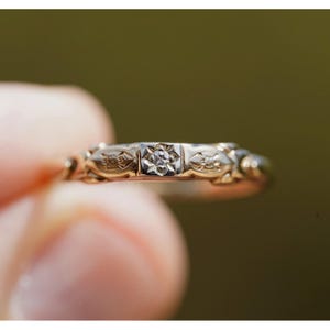 May include: A gold ring featuring a central square setting with a small, clear, round-cut gemstone. The band is decorated with an engraved, repeating floral or leaf-like pattern that extends across the visible surface of the metal ring.