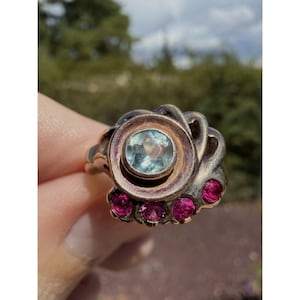 May include: A silver-toned ring featuring a central round light blue gemstone set within a circular frame, accented by four small round vibrant pink gemstones arranged in a curved row along the lower edge of the metal setting.