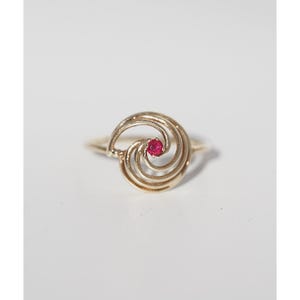 May include: A gold-colored ring with a swirling design. The ring features a small, round, red gemstone at the center of the swirl. The ring is set against a white background, highlighting its intricate details and elegant form.