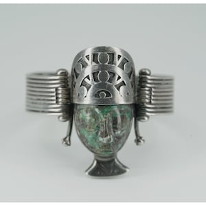 Op de afbeelding: Zilverkleurige ring met een centraal, gebeeldhouwd gezichtsontwerp in groen- en bruintinten. De ring heeft een decoratieve bovenkant met uitgesneden patronen en opgerolde banden aan weerszijden, wat een uniek, artistiek stuk creëert.