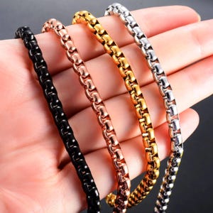 Puede incluir: Cuatro collares de cadena de metal en negro, oro rosa, oro y plata. Las cadenas se sostienen en una mano, mostrando sus diferentes colores y texturas. Las cadenas tienen un diseño de eslabones cuadrados redondeados.
