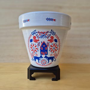 May include: A white ceramic plant pot with a hand-painted design in red and blue. The design features a blue house, squirrels, flowers, and stylized animals. The pot is on a small black stand.