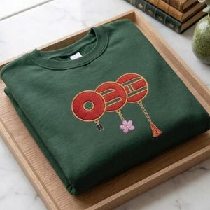 Pode incluir: Uma camisola verde-escura dobrada, com gola redonda, com um desenho bordado de três lanternas vermelhas com acabamentos dourados, uma flor rosa e uma borla. A camisola é exibida numa bandeja de madeira clara.