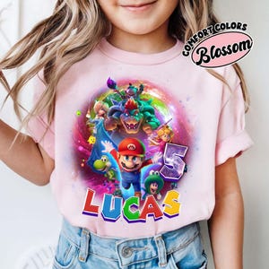 Puede incluir: Camiseta rosa claro con un gr&aacute;fico colorido de Mario y otros personajes de Super Mario Bros. El nombre "LUCAS" est&aacute; impreso en letras coloridas, con el n&uacute;mero "5" al lado. La camiseta tiene mangas cortas.