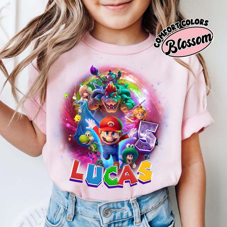 Puede incluir: Camiseta rosa claro con un gr&aacute;fico colorido de Mario y otros personajes de Super Mario Bros. El nombre "LUCAS" est&aacute; impreso en letras coloridas, con el n&uacute;mero "5" al lado. La camiseta tiene mangas cortas.