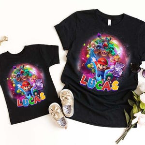 Puede incluir: Dos camisetas negras con un gr&aacute;fico colorido de Mario, Luigi y otros personajes. Las camisetas muestran el nombre "LUCAS" y el n&uacute;mero "5". La imagen incluye zapatos y flores, sobre un fondo blanco.