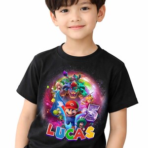 Puede incluir: Camiseta negra con un dise&ntilde;o colorido de Mario y otros personajes de Super Mario. El dise&ntilde;o incluye el n&uacute;mero "5" y el nombre "LUCAS". La camiseta la lleva un ni&ntilde;o peque&ntilde;o.