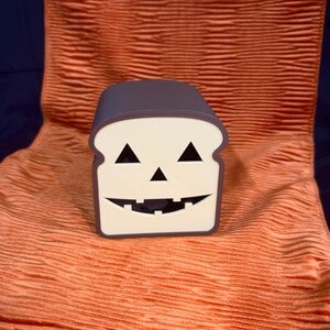 May include: A Halloween-themed bread-shaped container with a jack-o'-lantern face. The container has a white face with black triangle eyes and a jagged mouth. The top and sides are a dark gray color. It sits on an orange textured fabric.