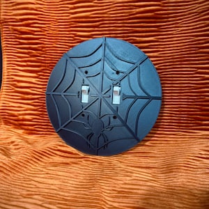 Könnte beinhalten: Eine schwarze, runde Lichtschalterabdeckung mit Spinnennetz-Design und einer Spinne in der Mitte. Sie hat zwei Lichtschalterwippen. Der Hintergrund ist orange und strukturiert.