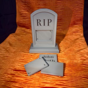 May include: A grey tombstone with the letters "RIP" at the top. Below the letters is a rectangular space. Two grey rectangular plaques are in front of the tombstone. The name "John Smith" is on one of the plaques. The objects are on an orange fabric.