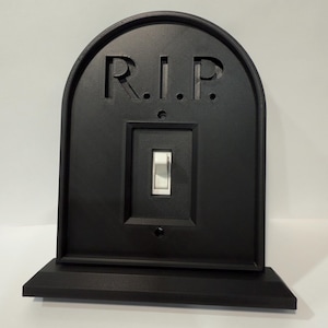 Könnte beinhalten: Eine schwarze Lichtschalterabdeckung in Form eines Grabsteins mit den Buchstaben "R.I.P." oben eingraviert. Der Schalter selbst ist weiß. Die Abdeckung hat eine rechteckige Basis.