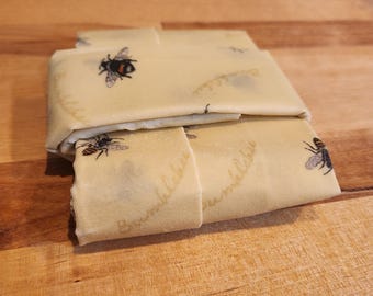 May include: Beeswax food wraps, folded and stacked, featuring a repeating pattern of bumblebees and the word "Bumblebee". The wraps are a light yellow color and appear to be made of a thin, flexible material.