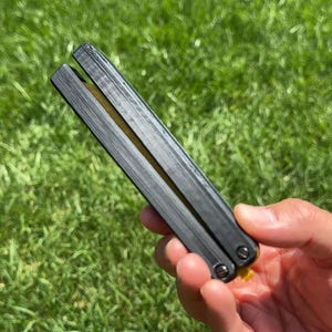 May include: A black butterfly knife with a textured handle is held against a backdrop of green grass. The knife is closed, showing the two halves of the handle joined together. The blade is not visible.