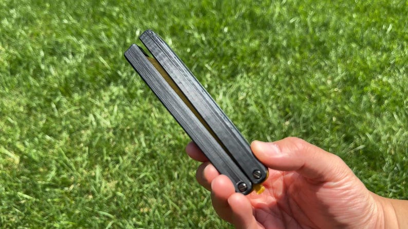 May include: A black butterfly knife with a textured handle is held against a backdrop of green grass. The knife is closed, showing the two halves of the handle joined together. The blade is not visible.