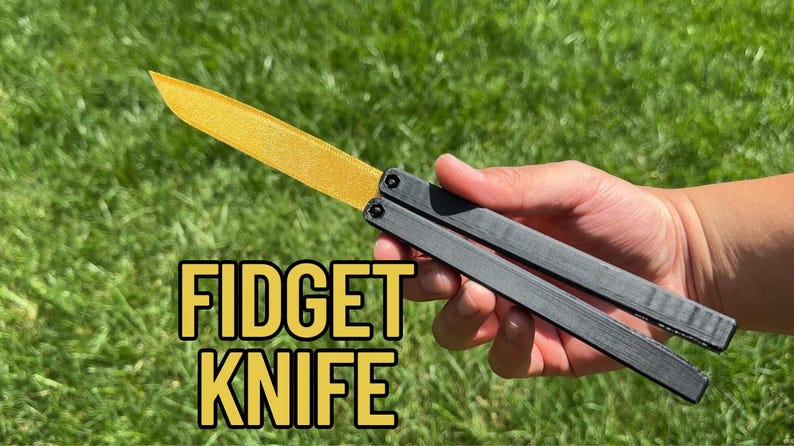 May include: A fidget knife with a gold-colored blade and black handle is held against a backdrop of green grass. The text "FIDGET KNIFE" is displayed in yellow block letters.