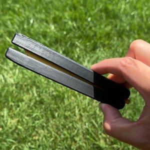 May include: A black butterfly knife with a gold-colored blade is held against a backdrop of green grass. The knife is open, revealing the blade. The handle is black and rectangular.