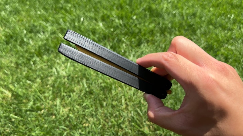 May include: A black butterfly knife with a gold-colored blade is held against a backdrop of green grass. The knife is open, revealing the blade. The handle is black and rectangular.