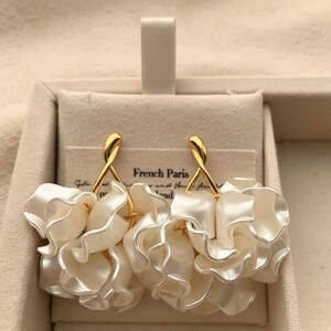 May include: Cream-coloured, petal-shaped earrings with gold-coloured accents. The earrings are displayed in a beige jewellery box. The text "French Paris" is visible on the card behind the earrings.