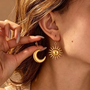 Puede incluir: Un par de pendientes colgantes de tono dorado con motivos celestiales. Un pendiente tiene forma de media luna lisa y pulida, mientras que el otro muestra un diseño de sol con una cara circular central y rayos ondulados radiantes.