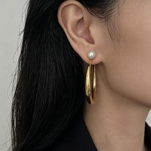Könnte beinhalten: Ein goldfarbener Creolen-Ohrring mit einem glatten, geschwungenen Metalldesign, das an einem einzelnen weißen, kugelförmigen Perlenstecker hängt. Der Ohrring wird am Ohrläppchen getragen und zeigt eine polierte Oberfläche.