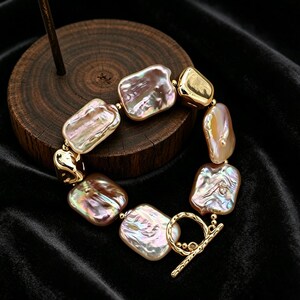 May include: A bracelet with large, irregularly shaped, iridescent pearls in pink, gold, and brown hues. The pearls are linked by gold-coloured metal and a toggle clasp. The bracelet is displayed on a dark, textured surface.