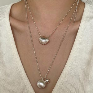 Op de afbeelding: Een gelaagde zilveren ketting set bestaande uit twee fijne balletjeskettingen. De kortere ketting heeft een kleine, geribbelde schelphanger, terwijl de langere ketting is voorzien van een grotere, gladde ronde hanger en een kleine zilveren kraal.