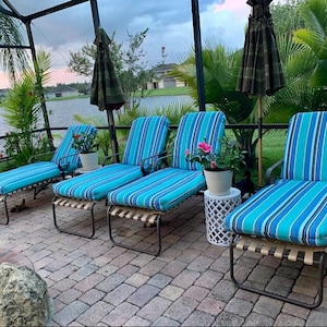 May include: Four blue and white striped lounge chairs with metal frames are arranged on a brick patio. The chairs are facing a body of water with a palm tree and a house in the background.