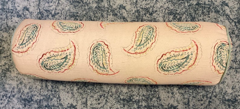 May include: A cylindrical bolster pillow featuring a light beige fabric base with a repeating paisley pattern in muted green and red tones. The ends of the bolster are finished with a solid light green piping trim along the circular edges.