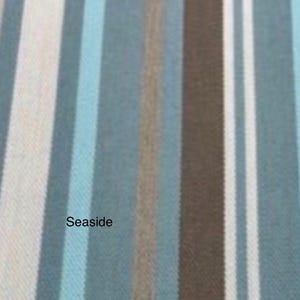 May include: A woven fabric featuring a vertical striped pattern in shades of light blue, dark blue, brown, and white. The word Seaside is printed in black text on the left side of the fabric, which displays a visible textile weave texture.
