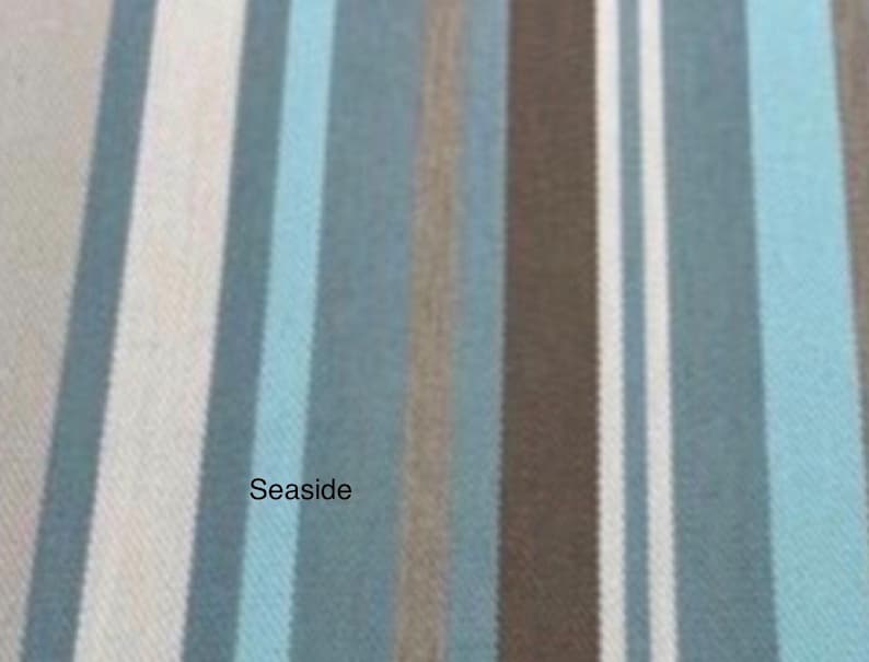 May include: A woven fabric featuring a vertical striped pattern in shades of light blue, dark blue, brown, and white. The word Seaside is printed in black text on the left side of the fabric, which displays a visible textile weave texture.