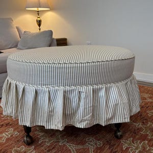 Peut inclure: Un pouf rond avec un dessus ray&eacute; et une jupe pliss&eacute;e. Le tissu pr&eacute;sente des rayures verticales blanches et gris-vert. Le pouf repose sur des pieds en bois fonc&eacute; avec des roulettes et est pos&eacute; sur un tapis &agrave; motifs.