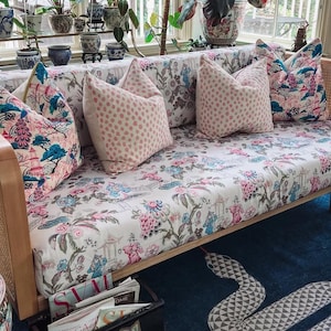 May include: A light wood sofa with cane side panels, upholstered in a white fabric featuring a multi-colored floral and pagoda pattern. The sofa includes two patterned throw pillows and two solid light pink polka-dot throw pillows.