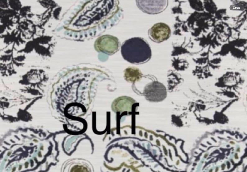 May include: A fabric pattern featuring a white background with black floral motifs, blue and green paisley shapes, and scattered dark blue and olive green circles. The word Surf is printed in black, bold, sans-serif capital letters across the center.