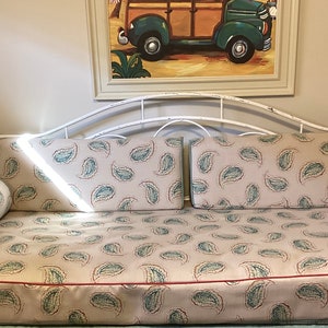May include: A white metal daybed frame featuring a curved backrest and side rails. The daybed includes a rectangular mattress and two cylindrical bolster pillows, all upholstered in a light gray fabric with a repeating teal and red paisley pattern.