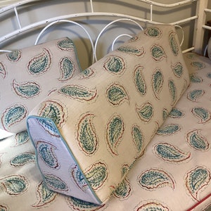 May include: A wedge-shaped bolster pillow and a rectangular pillow featuring a beige fabric base with a repeating teal and red paisley pattern. The wedge pillow includes light blue piping along the edges, while the flat pillow has red piping.