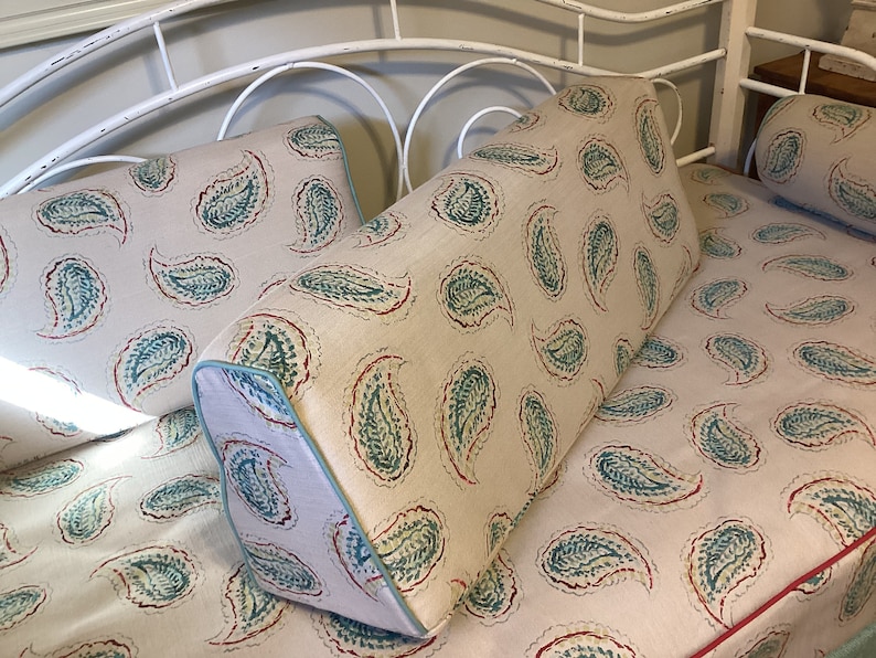 May include: A wedge-shaped bolster pillow and a rectangular pillow featuring a beige fabric base with a repeating teal and red paisley pattern. The wedge pillow includes light blue piping along the edges, while the flat pillow has red piping.