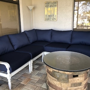 May include: A white sectional sofa with blue cushions sits on a patio with a stone fire pit table in the center. The sofa is made of wood and has a white painted finish. The fire pit table has a glass top and a stone base. The patio is made of brick pavers.