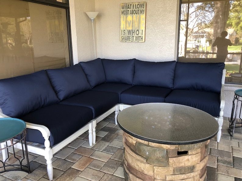 May include: A white sectional sofa with blue cushions sits on a patio with a stone fire pit table in the center. The sofa is made of wood and has a white painted finish. The fire pit table has a glass top and a stone base. The patio is made of brick pavers.