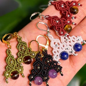 May include: A hand displays five pairs of macrame earrings in various colors. The earrings feature intricate knotwork and are adorned with gemstones in shades of brown, purple, and blue. The earrings have gold and silver-colored ear wires.