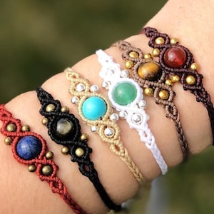 Può includere: Una collezione di braccialetti in macramè in vari colori, ognuno con una gemma centrale. I braccialetti includono cordoncini rossi, neri, bianchi e marroni, con gemme blu, verdi e di altri colori. Sono presenti anche piccole perline color oro.